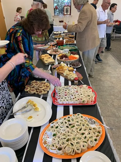 People serve themselves food from a buffet table covered with assorted appetizers, finger foods, and plates at a gathering.