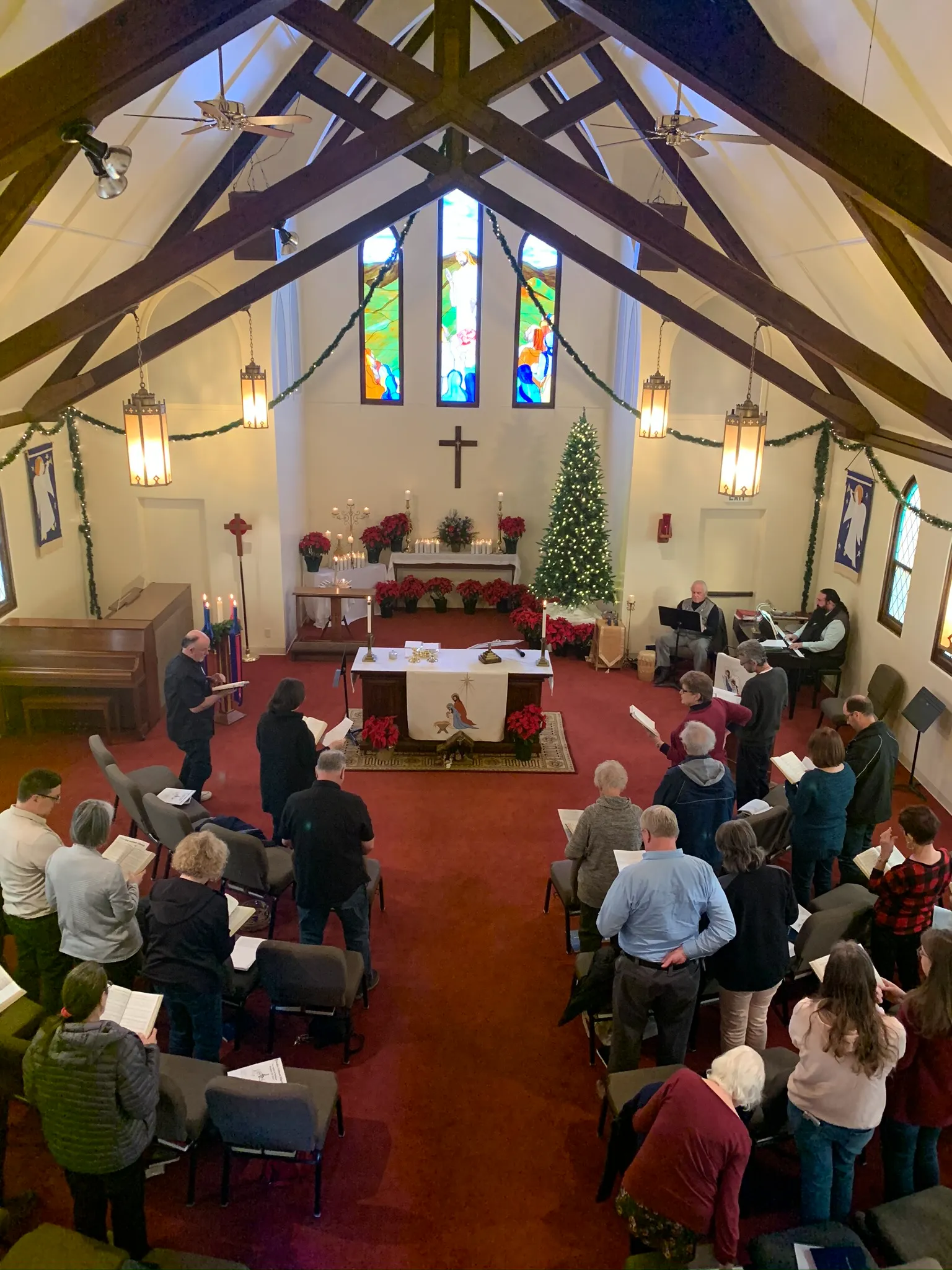 A congregation stands and sings inside a decorated church with stained glass windows, a Christmas tree, and an altar at the front.