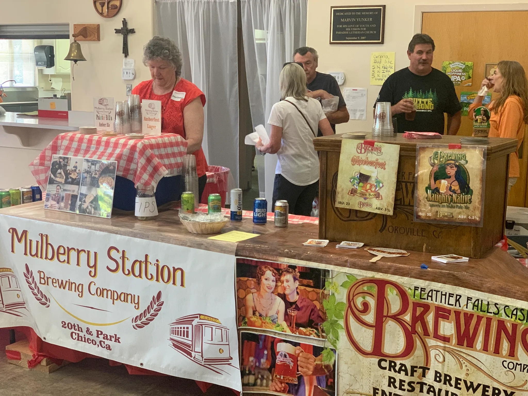 People stand behind and in front of tables displaying Mulberry Station and Feather Falls Brewing Company products at an indoor event. Signs, cans, and tasting cups are visible.
