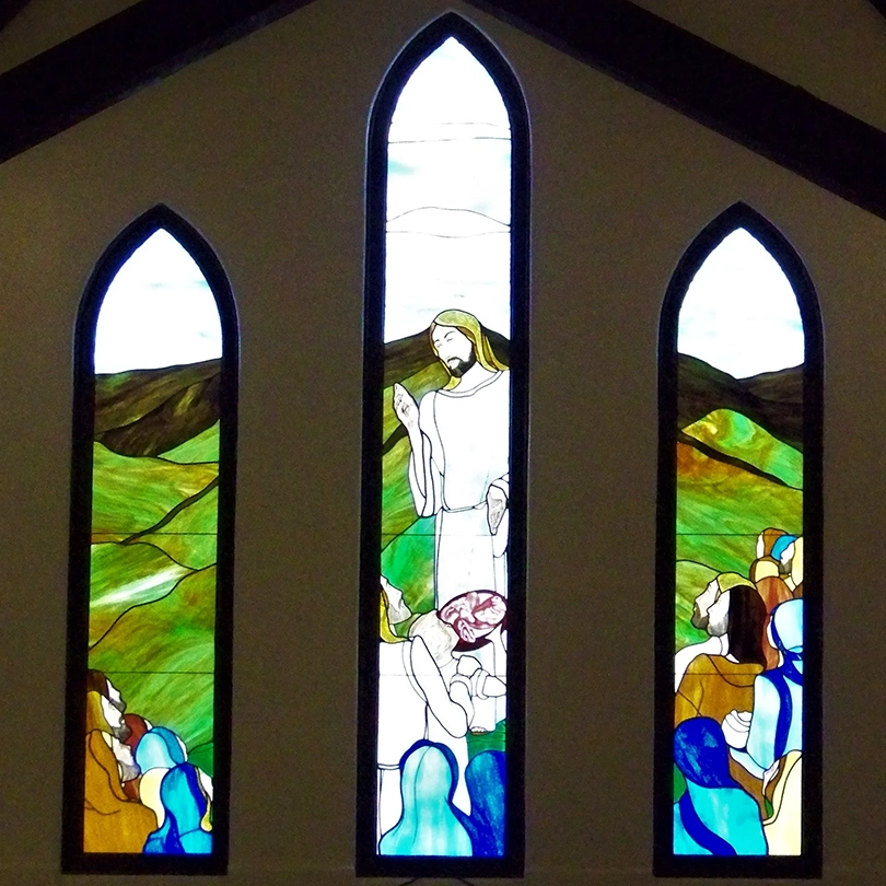 Stained glass window showing a robed figure teaching a seated crowd against a background of green hills and blue sky.