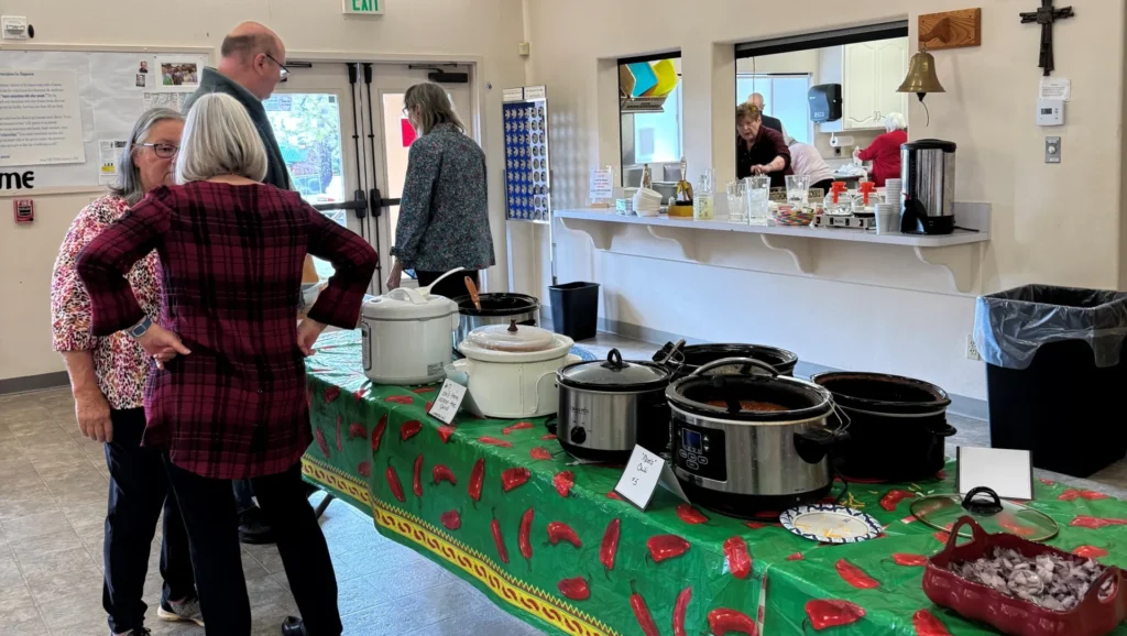 A group of people stands near a table with several slow cookers and labeled dishes at the 2026 Chili Cookoff, a lively community event in a hall featuring a delicious buffet setup.
