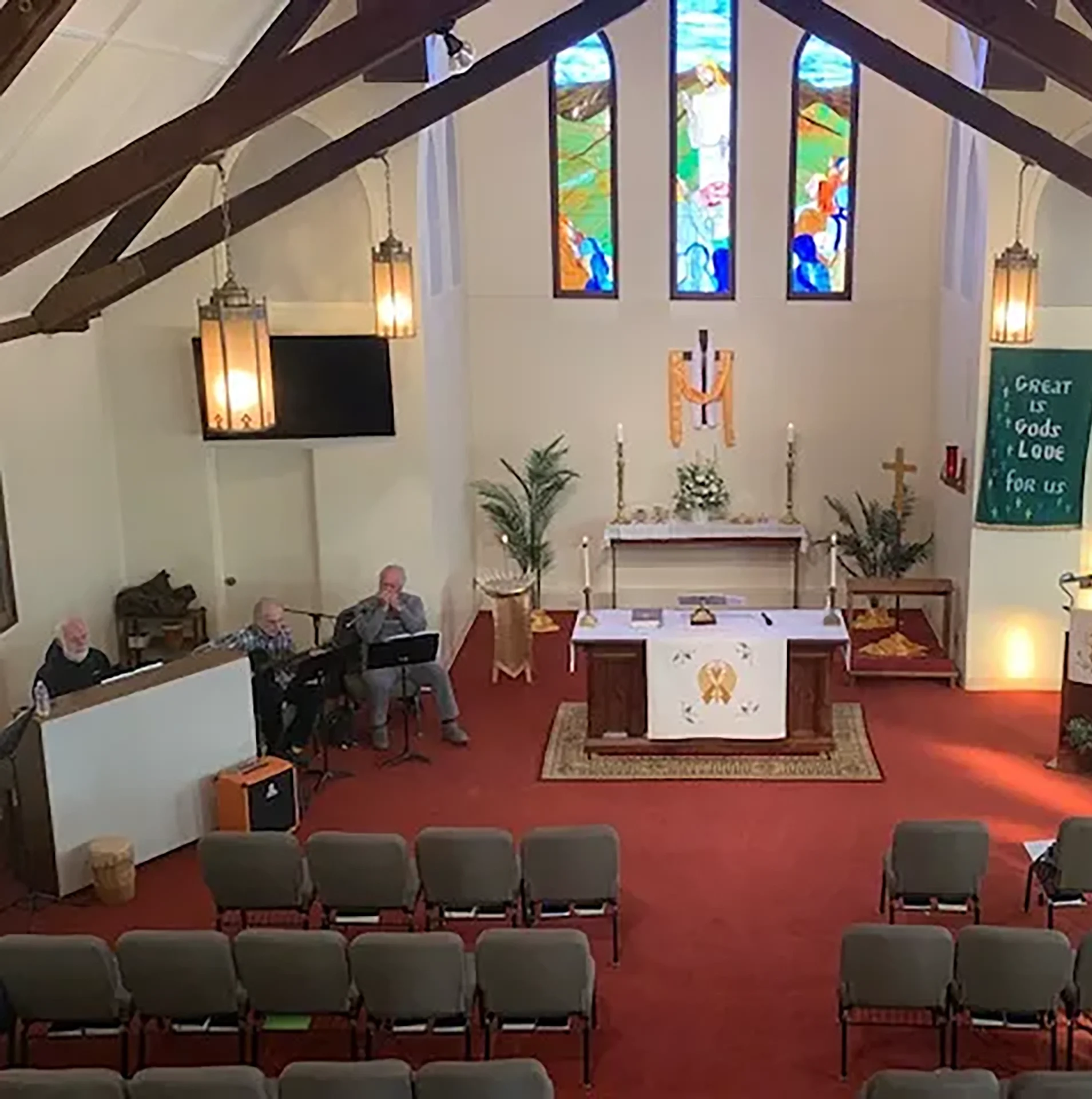 Interior of a small church with rows of chairs, altar at the front, musicians to the left, stained glass windows, and a banner reading, "Great is God's love for us.